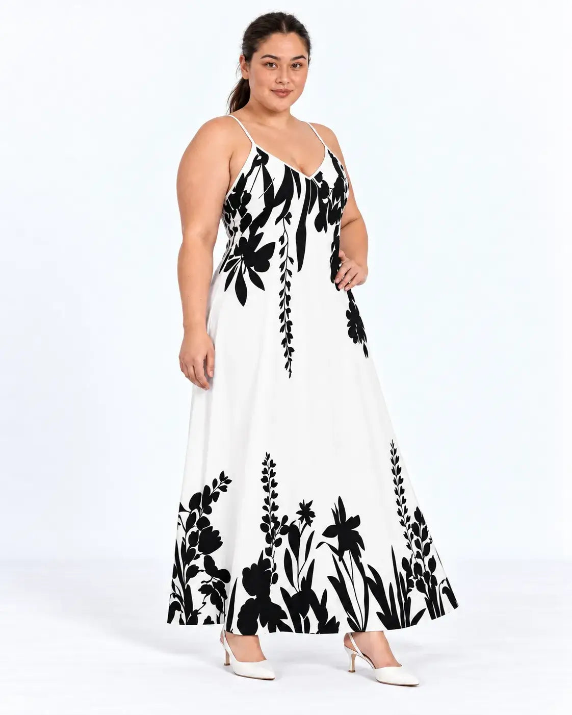 Modelo em pose de três quartos com vestido longo plus size branco, com alças finas e flores e folhagens pretas.