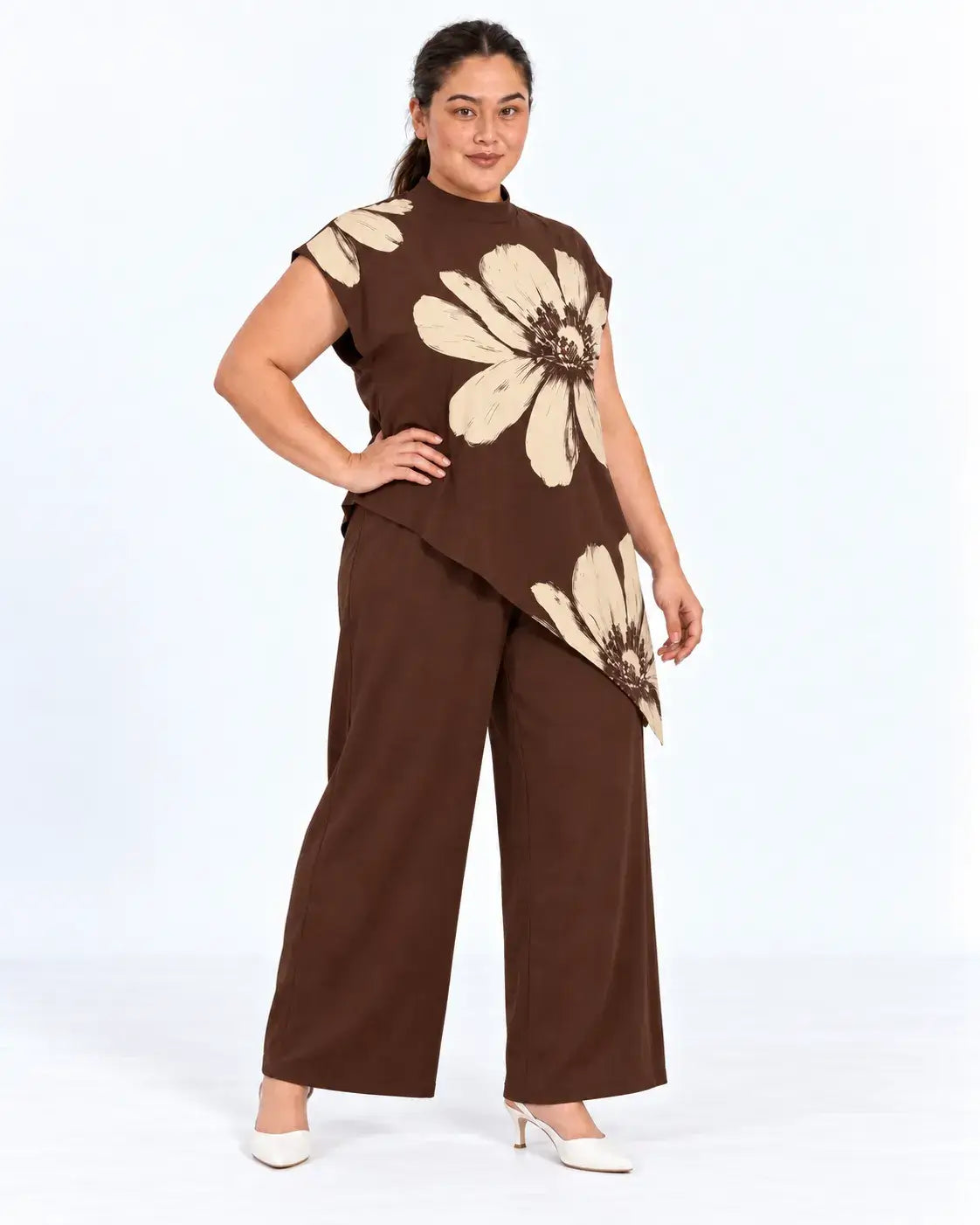 Modelo em pose de três quartos com blusa plus size floral castanha e bege, gola alta e barra assimétrica, mão na cintura.
