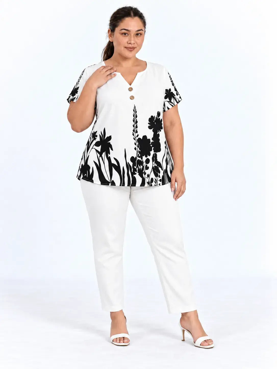 Modelo de frente usando blusa plus size floral preta e branca, com a mão junto ao decote e calça branca.