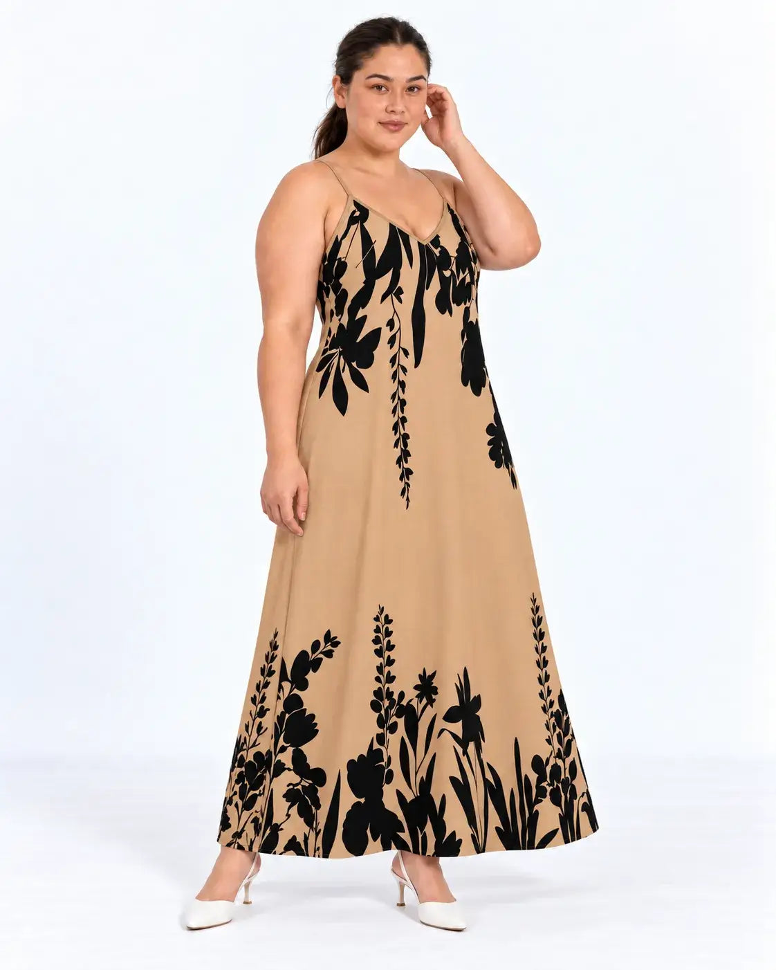 Modelo em pose de três quartos com vestido longo plus size marrom-claro e estampa botânica preta, com a mão no cabelo.